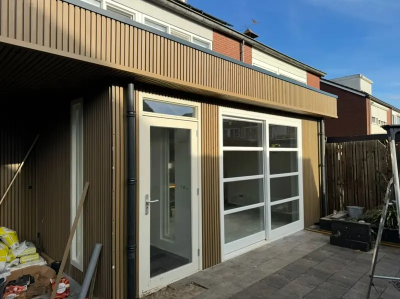 Opgeleverde uitbouw in Breda vanuit de tuin: verticale gevelbekleding, witte kozijnen met tuindeur en schuifpui, en een nieuw bestraat terras